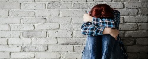 woman sitting against brick wall while hugging her knees with her head down